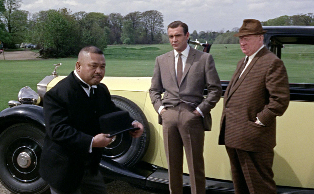 Oddjob (Harold Sakata) prepares to demonstrate his infamous hat trick to James Bond (Sean Connery) as Auric Goldfinger (Gert Fröbe) looks on.